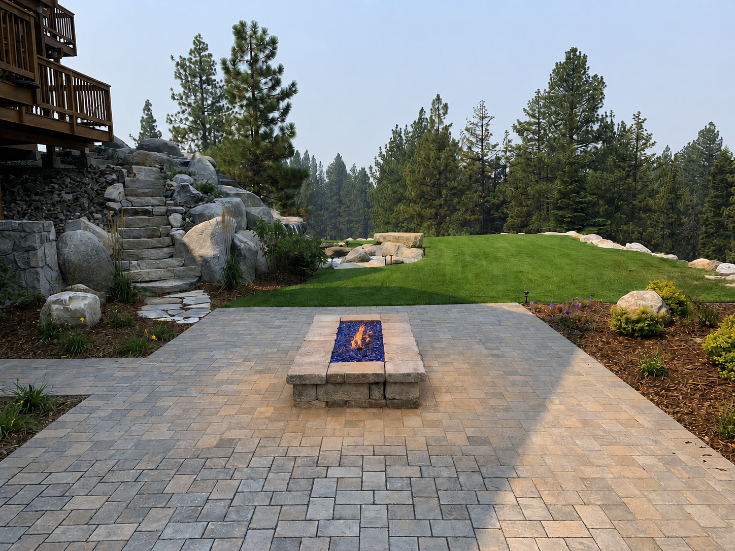 Paver patio with built-in fire feature, stone steps, lawn, boulders, and Tahoe pines