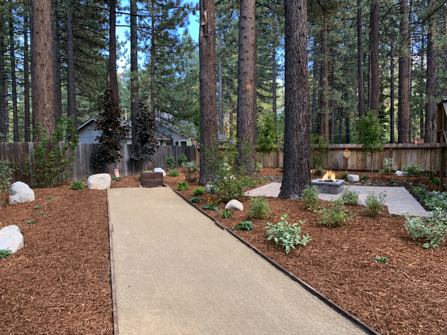 Freshly mulched Tahoe backyard with concrete pathway, granite boulders, planting beds, pine trees, and fire pit patio